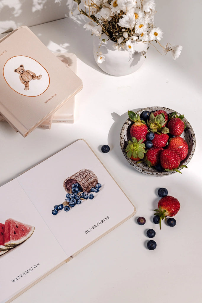 An artistically arranged photo featuring Our Little Library Book Box Set with watercolor illustrations of fruits, a bowl of strawberries and blueberries, and a floral arrangement, under soft lighting creates an inviting scene for introducing baby vocabulary.