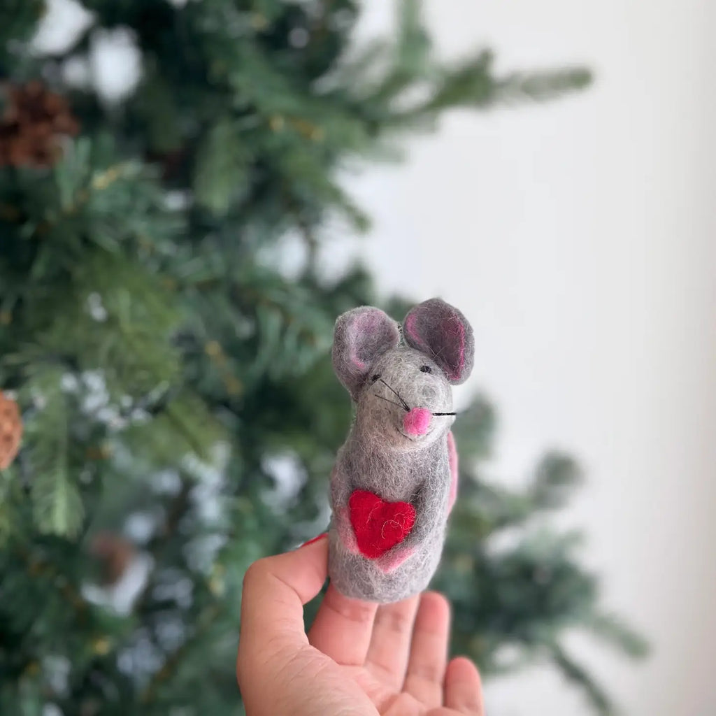 A "Mouse Holding A Heart Finger Puppet" ornament, handmade in Nepal with pink ears, a pink nose, and whiskers, is held in a person's hand against the backdrop of a green Christmas tree. The needle felted mouse proudly displays a red heart on its chest.