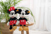 Two plush toys of Minnie Mouse and Mickey Mouse are sitting on a wooden stool. The Minnie Mouse toy, identified as the 12-inch Steiff Disney's Minnie Mouse Stuffed Plush Toy, is dressed in a red dress with white polka dots and a matching bow. She complements Mickey, who is wearing his classic red shorts with white buttons. They are positioned in front of a blurred green plant.