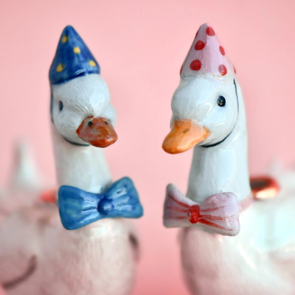 Two collectible keepsake Pink Goose Cake Toppers are adorned with colorful party hats and bow ties. The goose on the left wears a blue hat with yellow spots and a matching bow tie; the goose on the right has a pink hat with red spots and a matching bow tie. The hand-painted background is pink.