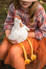 A child wearing a plaid shirt, orange tulle skirt, and a hat is sitting outdoors in a grassy area, holding the Charlie the Chicken Stuffed Animal—a soft toy with long yellow legs and a white body.