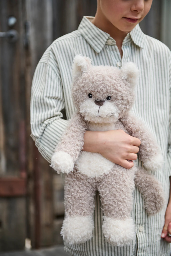 A child wearing a striped shirt holds a fluffy beige teddy bear with white patches, near a wooden fence. Checkers the Cat Stuffed Animal lounges nearby, creating an idyllic farm life scene.