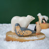 Handmade Wooden Penguins, including a polar bear, a penguin, and a seal, placed on a wooden platform with artificial snow. Two other figurines, one resembling a fox and another bird, are in the background against a dark green backdrop. These handcrafted linden wood toys are handmade in Serbia.