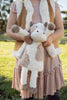 A child in a pink dress and brown vest is holding Clover The Cow Stuffed Animal, which has white and light brown fur. In the background, a sunny day outdoors is depicted with a grassy field and trees.