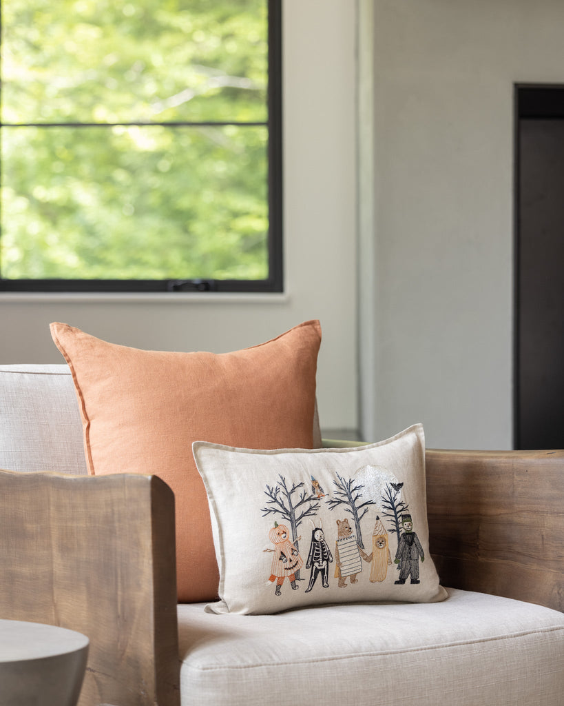 A cozy corner of a living room with a beige sofa adorned with a Full Moon Masquerade Pillow and an illustrated pillow, set against a window overlooking green trees.