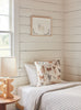 A cozy bedroom corner featuring a bed with a Coral & Tusk Dogs and Toys Pillow and a checkered blanket, beside a wooden nightstand with a lamp, all against a white paneled wall with a framed dog illustration.