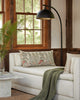 A cozy corner in a room with a white sofa adorned with a Coral & Tusk Mushrooms and Ferns Lumbar Pillow, a green throw, a small round table, and a tall floor lamp beside large wooden-framed.