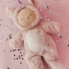 An Olli Ella Cozy Dinkum Doll Unicorn - Sugarpie, dressed in a fuzzy pink unicorn outfit, lies on a soft pink surface sprinkled with small silver star confetti.