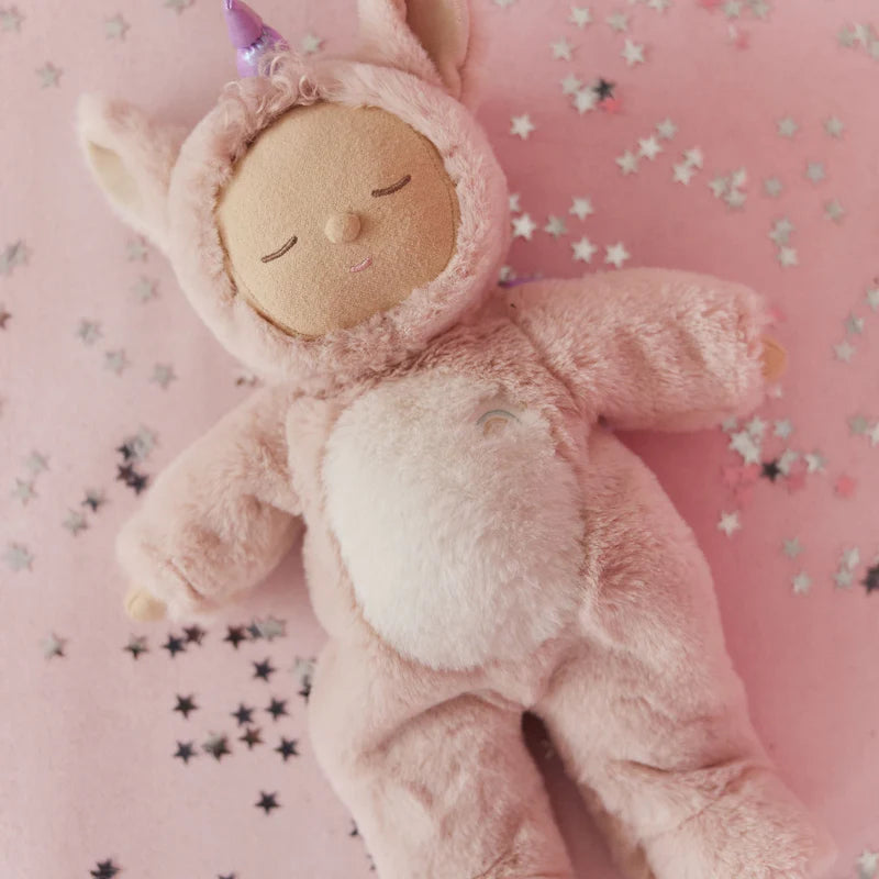 An Olli Ella Cozy Dinkum Doll Unicorn - Sugarpie, dressed in a fuzzy pink unicorn outfit, lies on a soft pink surface sprinkled with small silver star confetti.