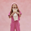 A child with long curly hair, wearing a pink beanie and outfit, laughs while holding the Olli Ella Cozy Dinkum Doll Unicorn - Sugarpie, featuring shimmering wings, against a pink background.