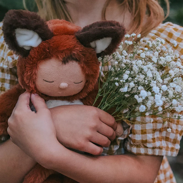 A child in a checkered dress holds an Olli Ella Cozy Dinkum Doll - Fox along with a small bouquet of white flowers. The scene conveys a sense of warmth and gentleness, reminiscent of the charming designs from Olli Ella.