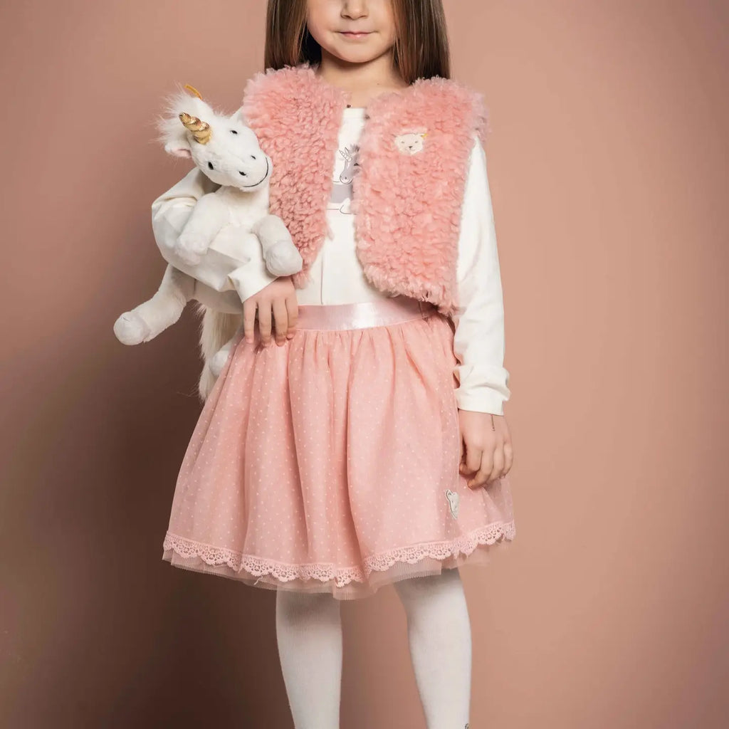 A girl in a pink skirt and white top holds a Steiff Unicorn Stuffed Animal, 11 Inches, standing against a soft beige background. She wears a cozy pink fur vest and white tights.