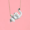 A delicate Gold Tiger Necklace featuring a small, playful white tiger pendant with black stripes, depicted hanging upside down, grasping the 24k gold plated steel chain with its paws. The hand-painted porcelain charm stands out beautifully against the solid pink background, highlighting the charming design.