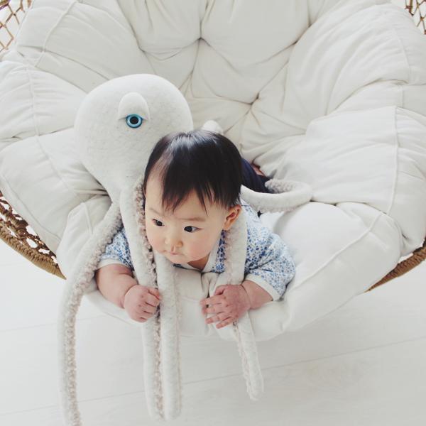 A baby lies on their stomach in a cozy, round, white cushioned chair, holding onto a BigStuffed Octopus - Big Albino. The baby looks curious and engaged as if exploring an underwater world, with the octopus toy's tentacles draped over them and the chair.