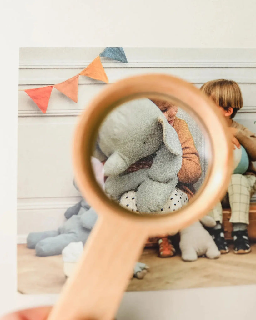 A Wooden Magnifying Glass focuses on a child holding a plush elephant, crafted from Beech Wood, with part of another child visible. The background shows a festive setting with a banner and other plush toys.