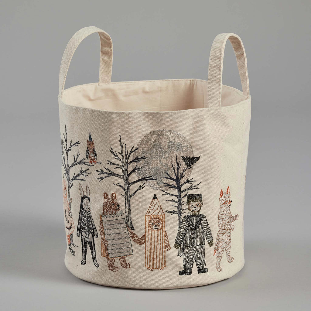 The Coral & Tusk Full Moon Masquerade Bucket is a beige fabric storage basket with two handles, featuring an embroidered design of children dressed in various animal and character costumes standing in a line among leafless trees. The background includes a large full moon and a flying bat, making it perfect as a Halloween candy bucket.