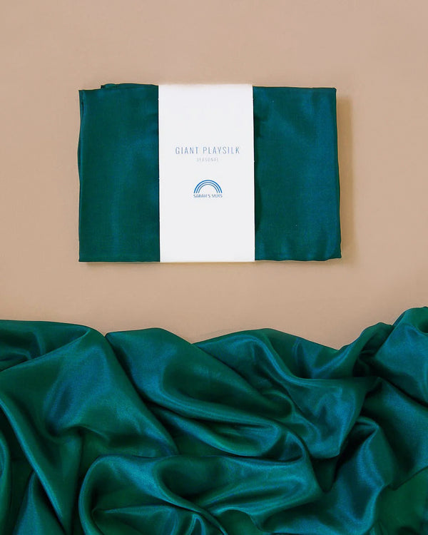 A neatly folded Giant Evergreen Playsilk from Sarah's Silks is displayed against a beige background, featuring a white label. Crafted from luxurious mulberry silk, the teal fabric drapes below, highlighting its smooth and shiny texture in vibrant jewel tones.