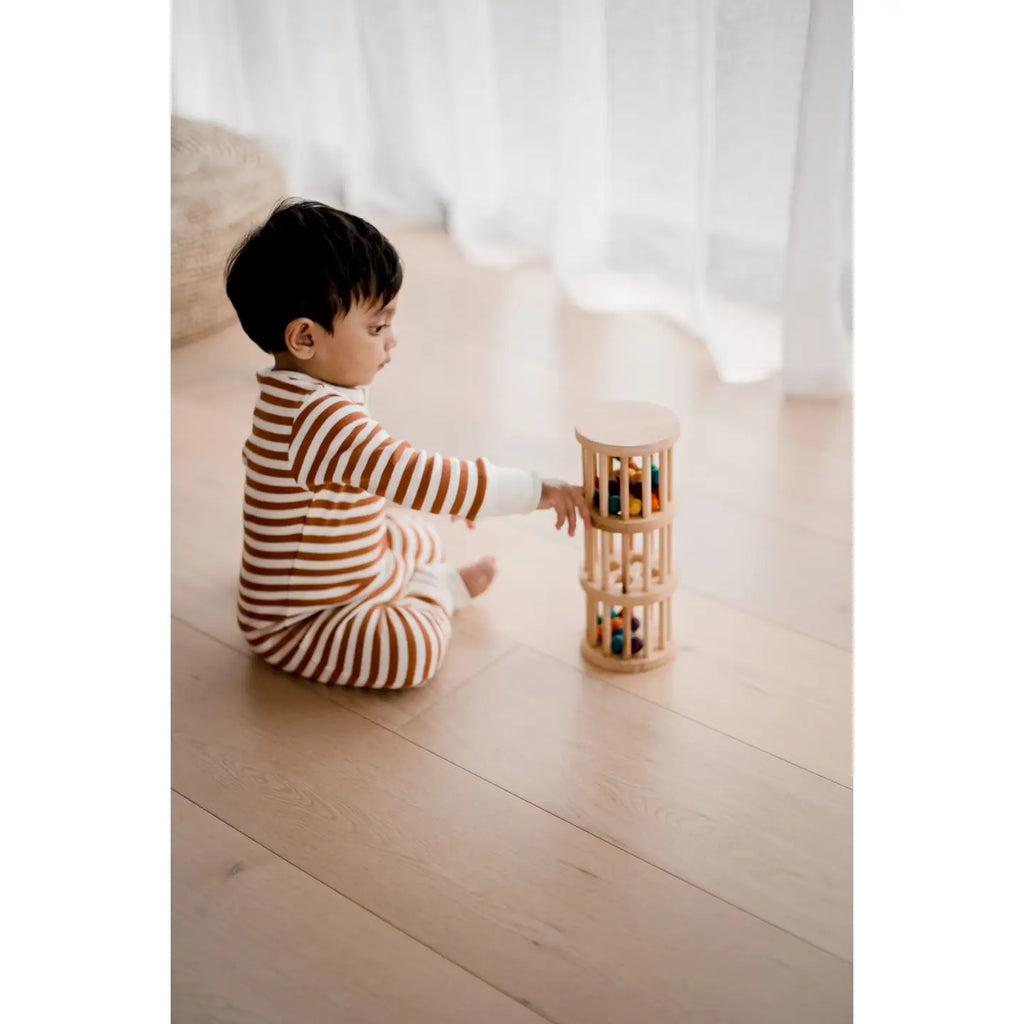 A young child in a brown and white striped onesie sits on a wooden floor near a window with sheer curtains, playing with the Wooden Rain Maker, a cylindrical wooden toy filled with colorful, non-toxic balls.