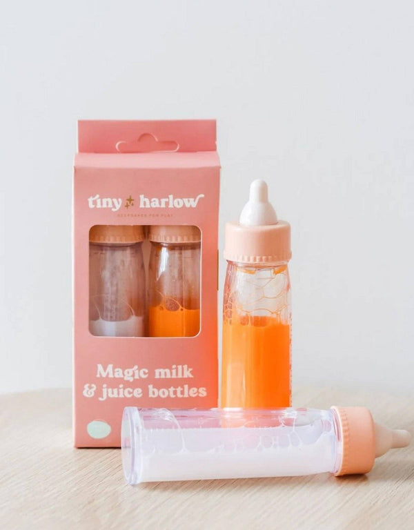 The Bottled Milk and Juice Set by Tiny Harlow is displayed with its packaging. One toy baby bottle is filled with orange liquid, and the other remains empty. The pink and white package features the label "Tiny Harlow Magic Milk & Juice Bottles." Perfect for doll feeding, this charming set is placed on a light wooden surface.