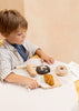 A young child sits at a table, joyfully engaged with the Wooden Bakery Set, which includes toy bread and pastries such as a loaf, donut, bagel, and croissant. The child carefully slices the croissant with a wooden knife while dressed in a striped shirt and navy blue t-shirt.