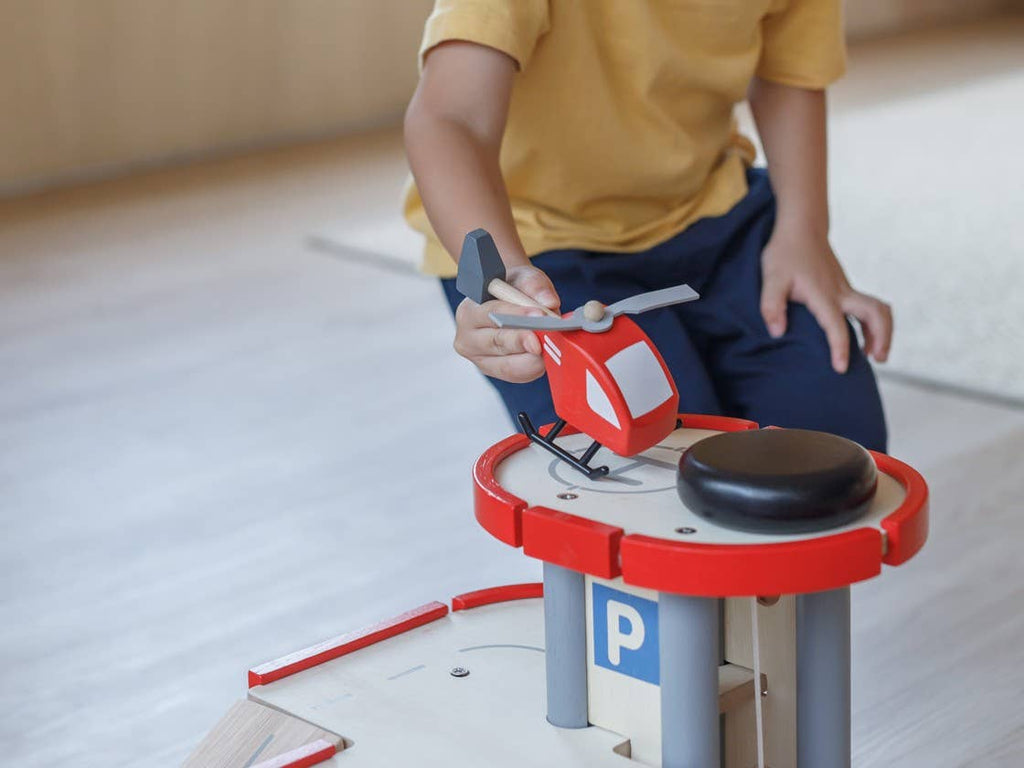 A child engages in pretend play with a red Helicopter, zooming it around a sustainably made wooden parking toy set.