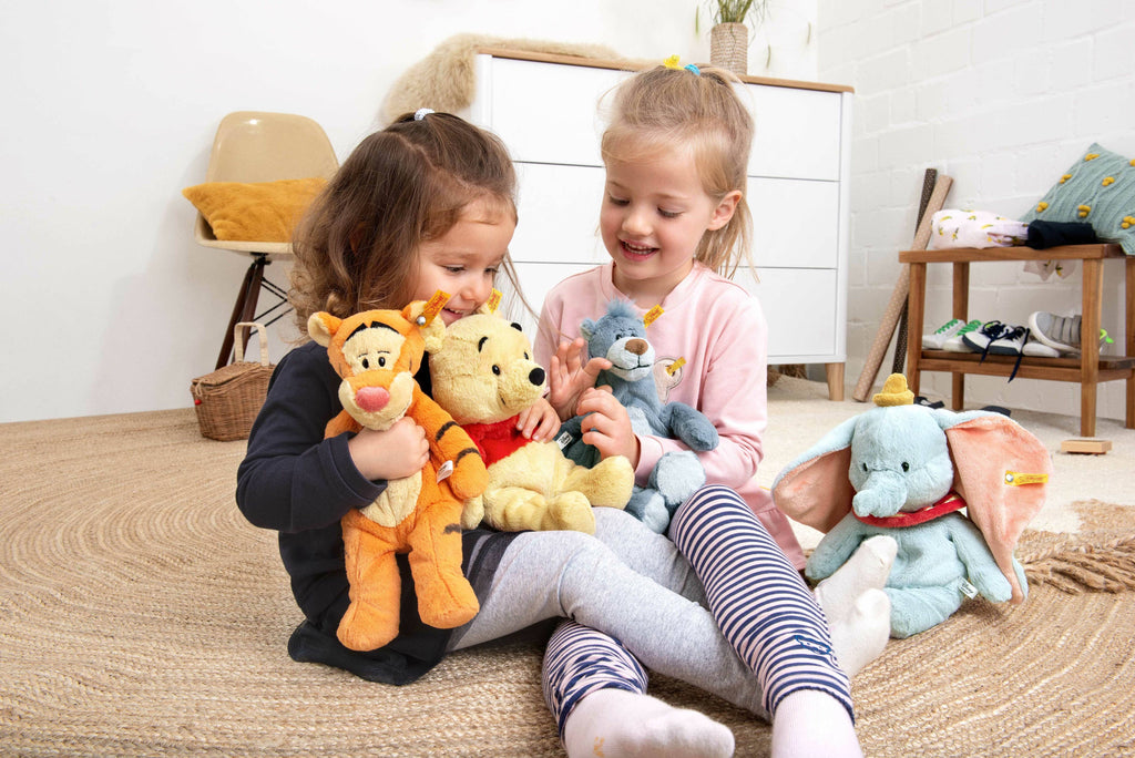 Two young girls sit on a beige carpet, grinning as they play with plush toys, including a 12-inch Steiff Disney's Tigger the Tiger stuffed plush toy, alongside Winnie the Pooh and Dumbo, in a cozy room with a chest of drawers in the background.