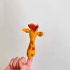 A hand is shown wearing a Giraffe Finger Puppet, which is yellow with brown spots and features small ears and a tuft of orange hair on top, mimicking a giraffe's head. The background is plain and white.