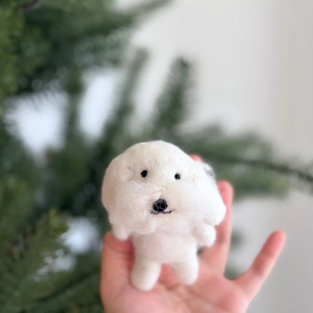 A hand is holding a small, plush Maltese Dog Finger Puppet made from 100% wool in front of a blurred background with green foliage. The needle-felted figurine has black eyes and a black nose, giving it a cute, fluffy appearance.