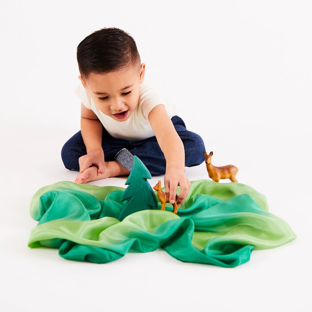 A child sits on the floor, immersed in imaginative play with a forest-themed toy set. Complete with Sarah's Silk Enchanted Playsilk - Forest serving as lush green grass, a small tree, and deer figurines, these open-ended toys captivate the child's curiosity and creativity.