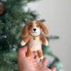 A hand is holding a small, handmade Cavalier King Charles Spaniel Dog Finger Puppet with brown and white fur and a cute expression. The finger puppet, featuring intricate needle felt details, is wearing a tiny bell on a black string around its neck. The background features a blurred out Christmas tree.