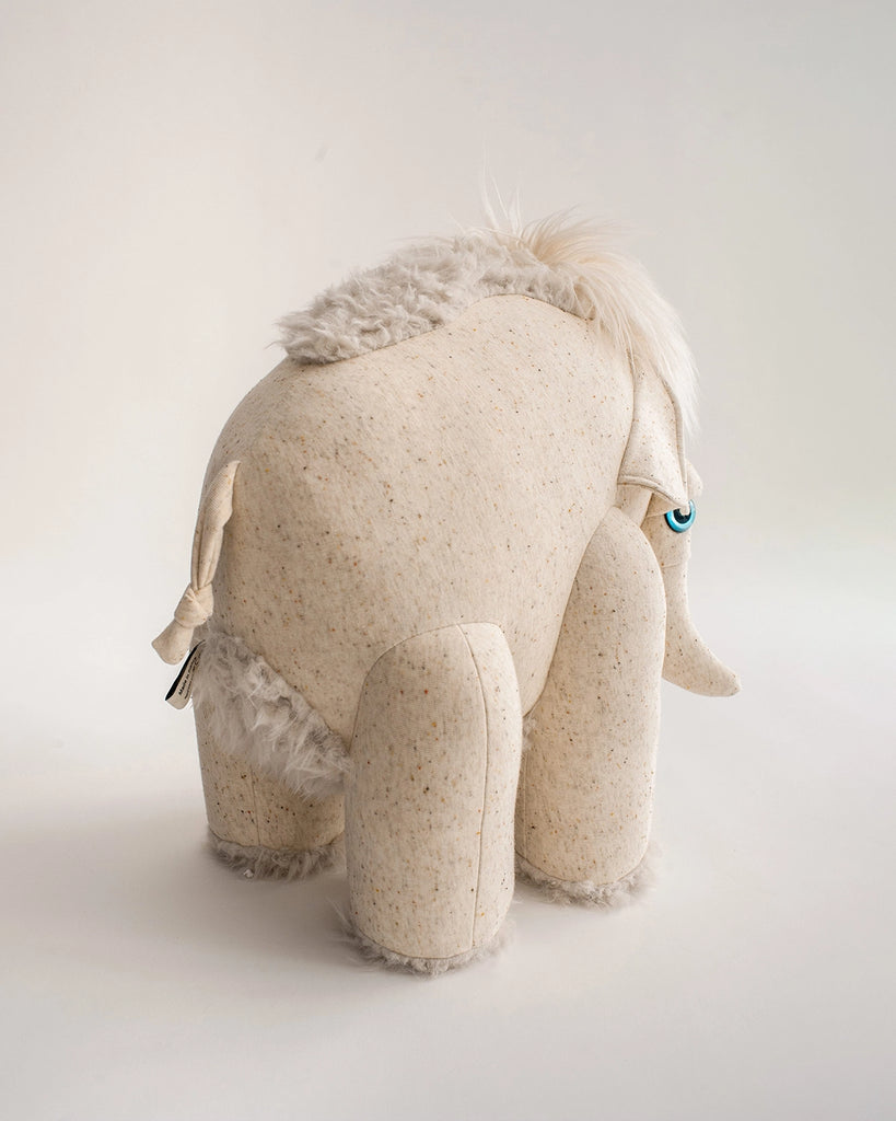 A soft toy resembling a stylized, anthropomorphic elephant stands on all fours. It has a textured cream body, fuzzy patches of fur on its back and head, a tufted tail, and a single visible blue eye with a playful expression. This BigStuffed Albino Mammoth Stuffed Animal is set against a plain light-colored background.