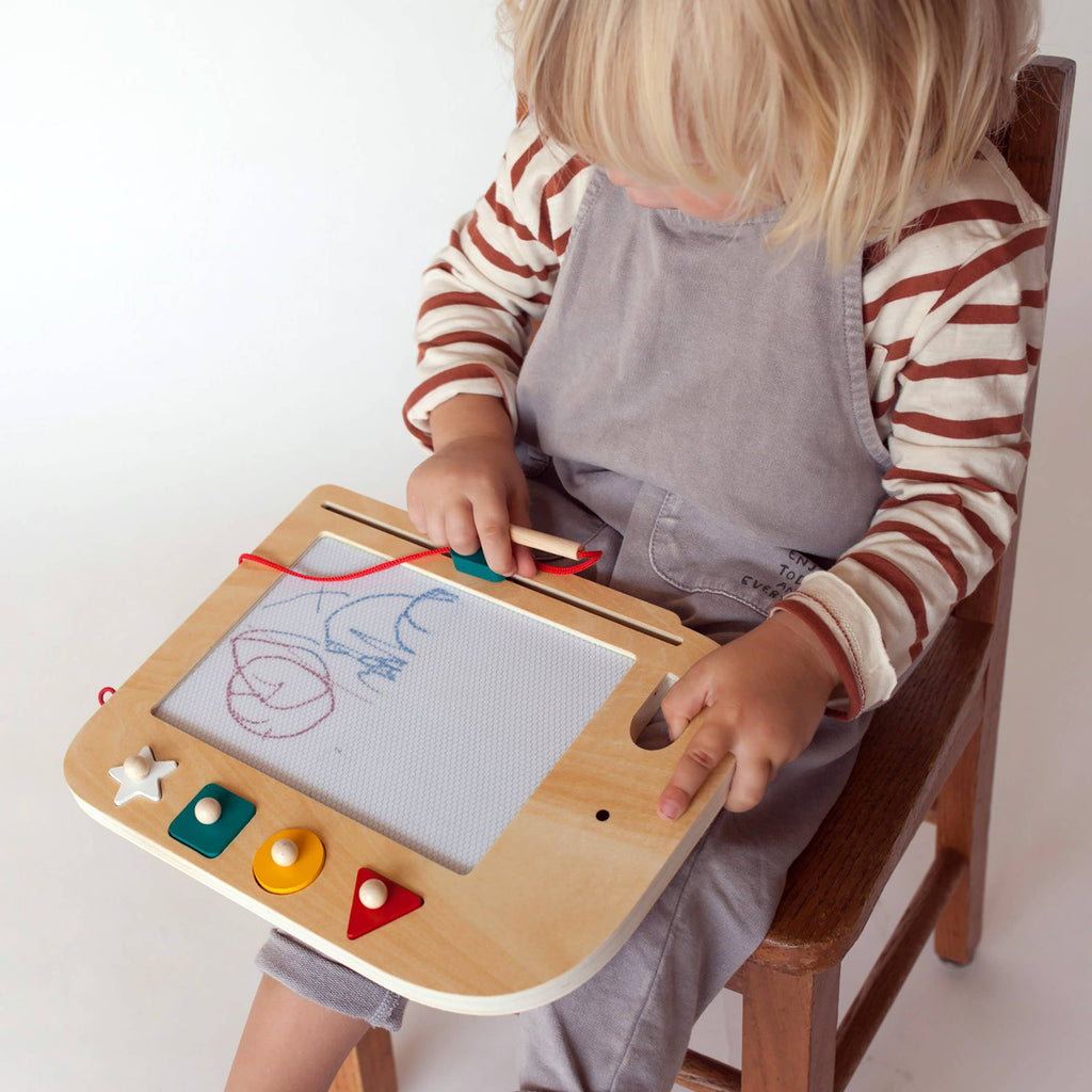 A child with blonde hair is seated on a wooden chair, wearing a gray apron over a striped shirt. They are using the Wooden Elephant Magic Drawing Board with colorful shape buttons, and there’s a magnetic pen as the child draws on the board.