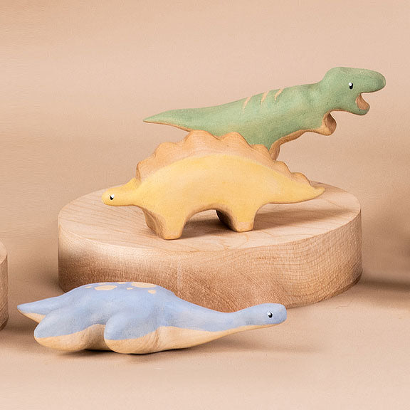 Three handmade wooden plesiosaurus dinosaur toys on a beige background. One resembling a t-rex stands upright, another shaped like a stegosaurus is on a wooden base, and a third resembling a br