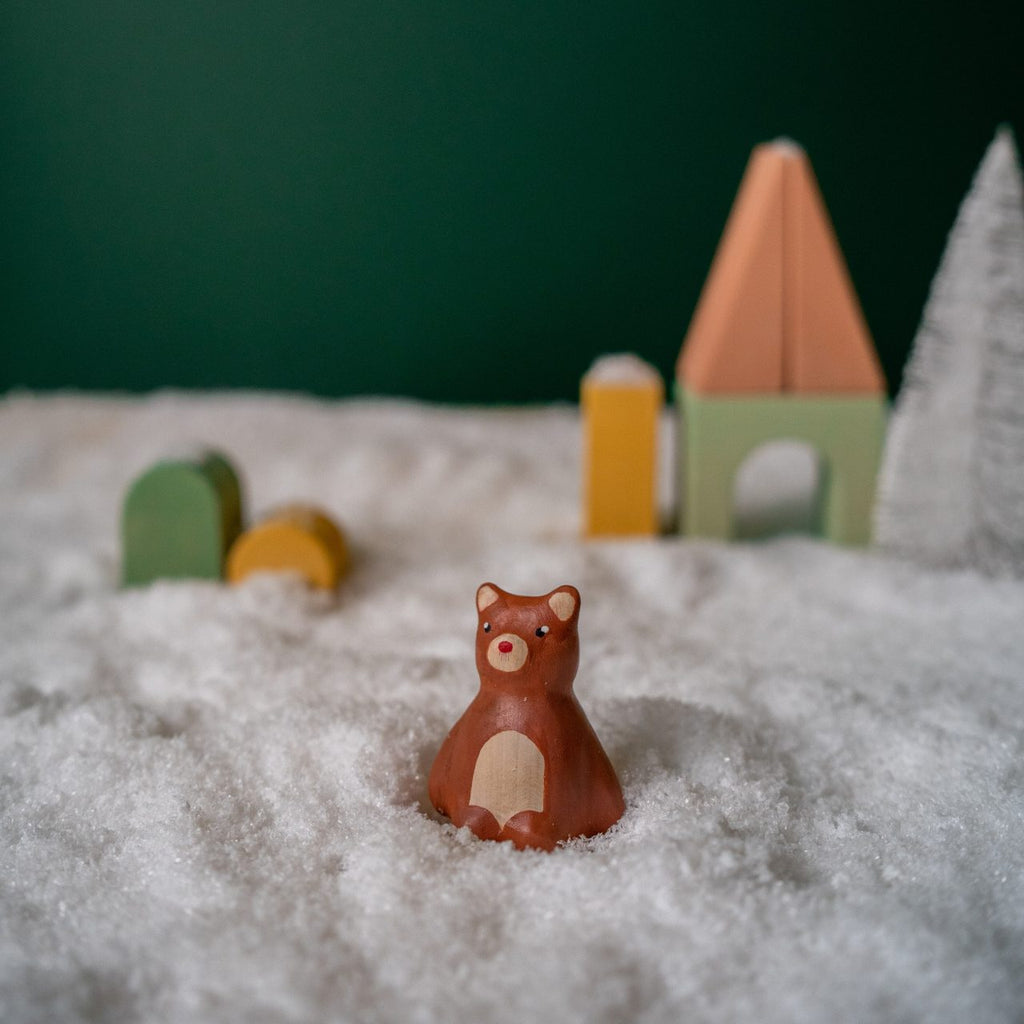 A delightful piece from the Handmade Wooden Christmas Characters Set, a small linden wood bear toy stands amidst soft, textured snow in a charming winter scene. In the background, pastel-painted abstract wooden shapes resembling trees and a house contrast beautifully against a dark green backdrop. These figures are part of the Christmas collection and are lovingly handcrafted with child-safe paint.