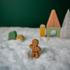 A small gingerbread man figurine stands on artificial snow in front of pastel-colored, handcrafted linden wood toys resembling houses and trees. A dark green backdrop contrasts the snowy scene, creating a whimsical winter landscape. This piece is part of our Handmade Wooden Christmas Characters Set.