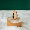 A Handmade Wooden Penguin stands on a cylindrical platform, handcrafted from linden wood and covered with a light dusting of non-toxic child-safe paint to mimic snow. Handmade in Serbia, the background is green, enhancing the snowy scene.