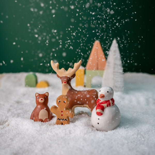 A festive scene featuring the Handmade Wooden Christmas Characters Set, with small carved figures of a reindeer, snowman, bear, and gingerbread person standing in artificial snow. In the background, there are miniature wooden trees and houses set against a dark green backdrop with falling snow. These child-safe painted figurines beautifully capture the spirit of Christmas.