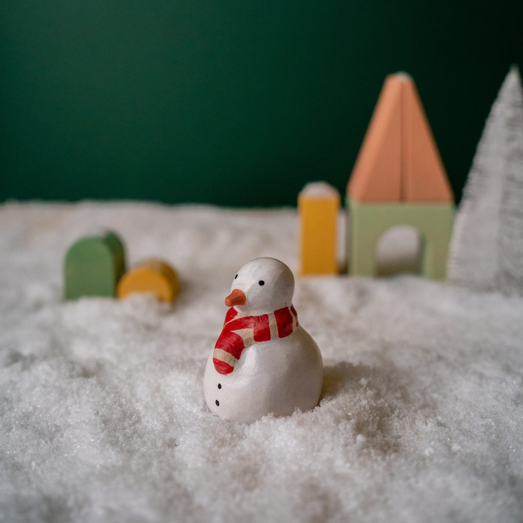 A small snowman figurine with an orange carrot nose and a red scarf from the Handmade Wooden Christmas Characters Set stands on a snow-covered surface. In the background, there are blurred wooden toy houses and trees, handcrafted from linden wood, painted in soft pastel colors with child-safe paint, set against a dark green backdrop.