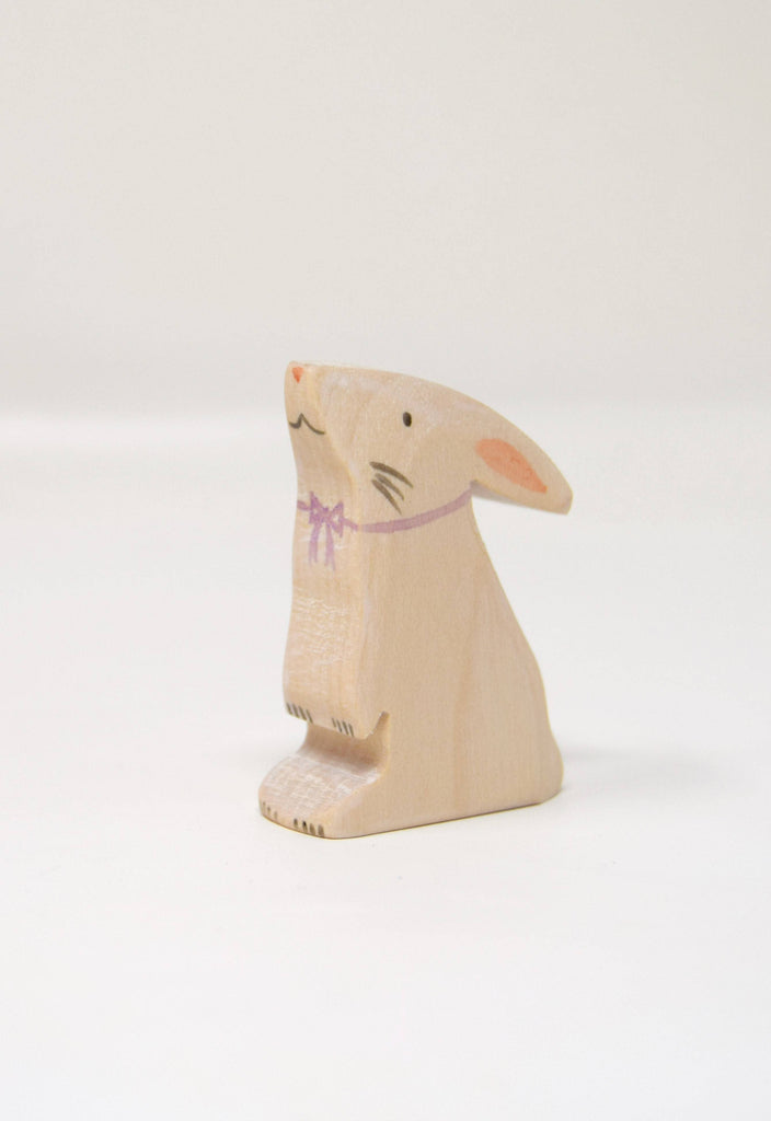 The Handmade Wooden Easter Bunny is a small, light figurine crafted from wood, featuring minimalistic hand-painted details and a simple pink bow around its neck, set against a plain white background.