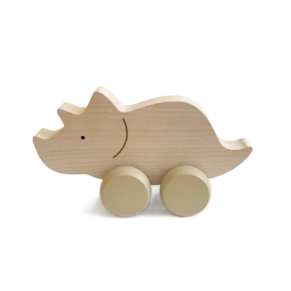 A Wooden Triceratops Dinosaur Push Toy, handmade in Poland, boasts simple carved details like a small eye and a smiling mouth. Equipped with four round wheels for rolling, this delightful toy promotes fine motor skills development and features a natural, light wood finish.