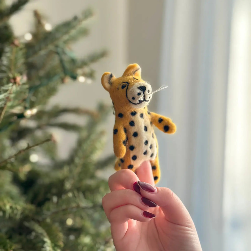 A hand with painted nails holds up a Cheetah Finger Puppet in front of a decorated Christmas tree with blurred lights in the background. The handmade toy, small and yellow with spots, adds a touch of whimsy to the festive scene.