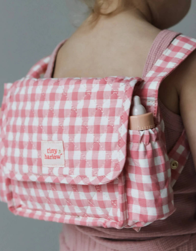 A child equipped with the Tiny Tummies Pink Gift Box from Tiny Harlow, showcasing doll essentials, carries a pink and white checkered backpack. This charming accessory is labeled "tiny harlow" and features a front flap and side pocket perfect for holding a small baby bottle. Dressed in a complementary pink outfit, the child looks poised for adventures.