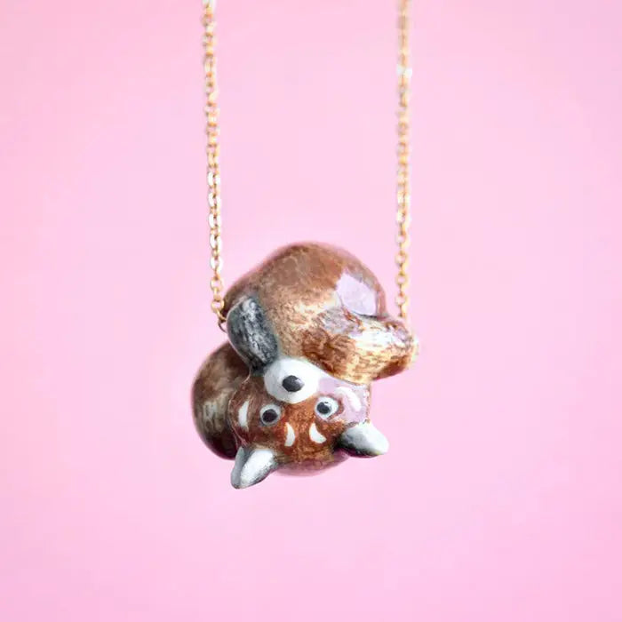 A necklace featuring a cute, tiny red panda pendant hanging upside down is pictured against a pink background. The red panda has wide eyes and pointed ears, and the pendant is attached to a delicate 24k gold plated chain.