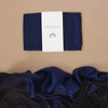 A piece of dark blue silk, folded and wrapped with a white label reading "Sarah's Silk Giant Midnight Playsilk," unfurls on a beige surface, inviting imaginative play with its smooth, rich texture. It's perfect for open-ended play adventures.