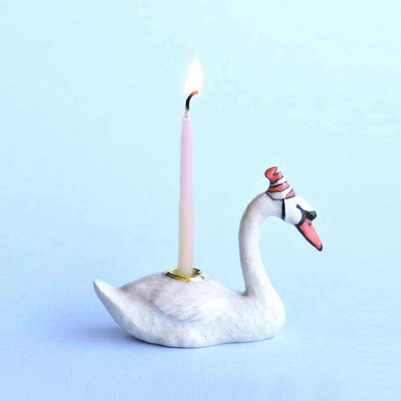 A decorative Swan Cake Topper shaped like a swan with a lit wick, placed against a soft blue background. The swan, a hand-painted porcelain piece, wears a small, red and white striped