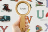 Sentence with replaced product name: A hand holding a Wooden Magnifying Glass over the word "tree" on a colorful educational poster featuring various illustrations and alphabets.