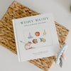 A children's board book titled "Wishy Washy: A Board Book of First Words and Colors" is placed in a woven basket, accompanied by a silicone teething ring. The book cover features illustrations.