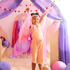 A young child stands inside a cozy, fabric-draped play fort, smiling widely and holding up a Sarah's Silk Blossom Garland Headband with joy. The fort is decorated with heart-shaped garlands and includes soft toys and colorful fabric hanging from the wooden frame, perfect for dress-up play adventures.