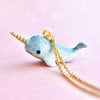 A small Narwhal Necklace with a gold chain. The hand-painted charm features a light blue narwhal with a spiral tusk and a cute smile, set against a soft pink background.