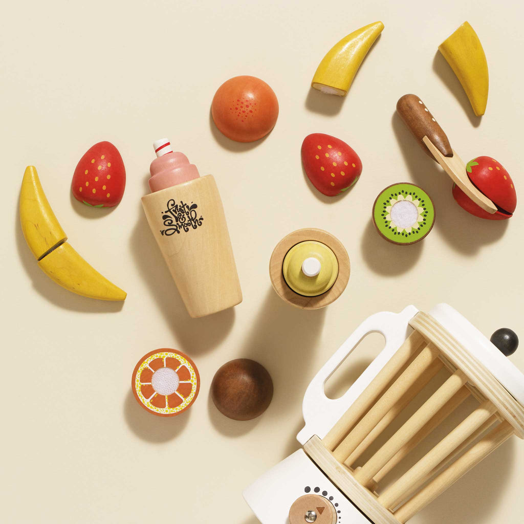 The Fruit & Smoothie Blender Set, featuring an assortment of sliceable wooden toy fruits such as bananas, strawberries, slices of kiwi, and oranges, is artfully arranged around a retro-style blender and a wooden bottle on a light background in an enchanting wooden toy kitchen setting.